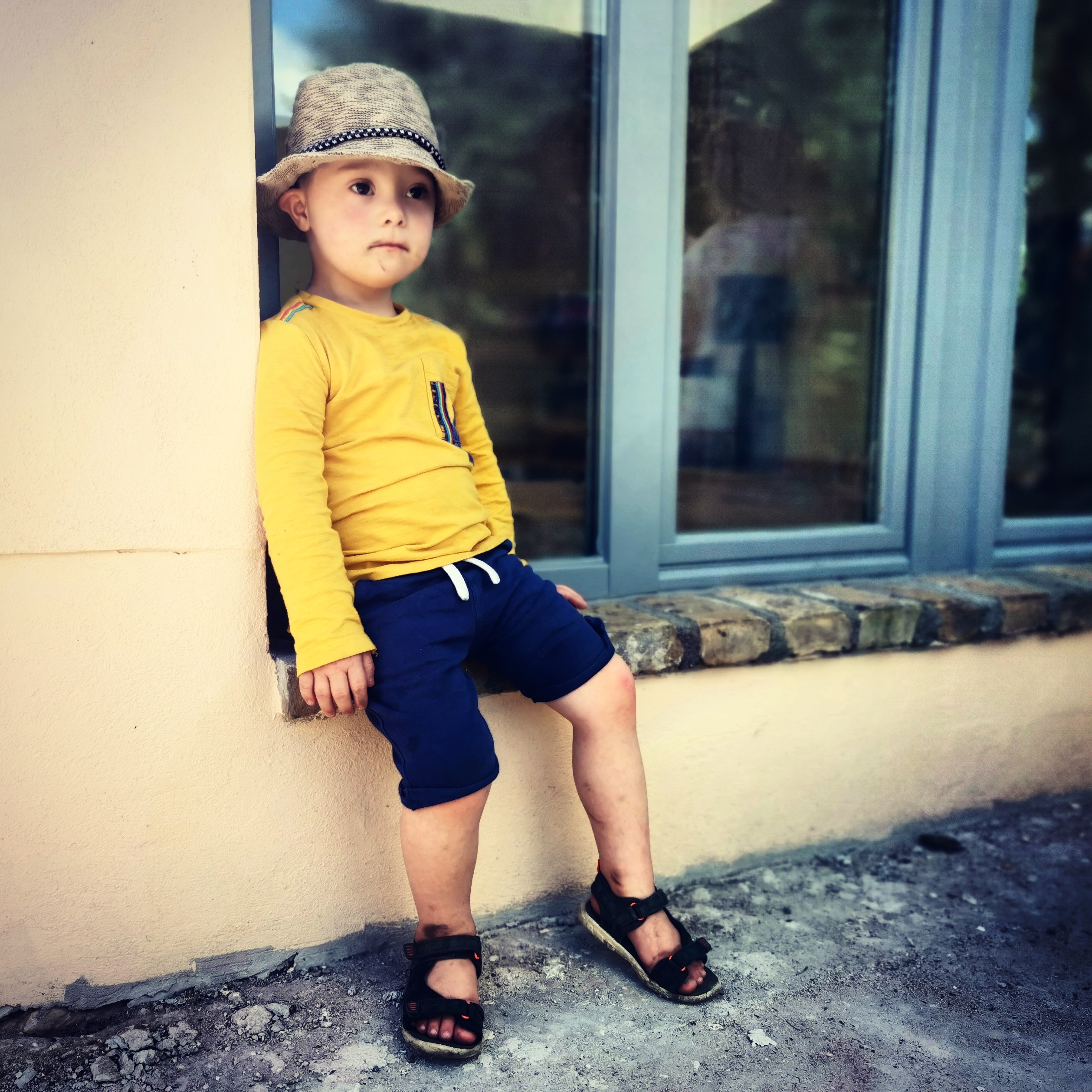 A little boy in blue shorts, a yellow tshirt and a hat sit on a window sill.