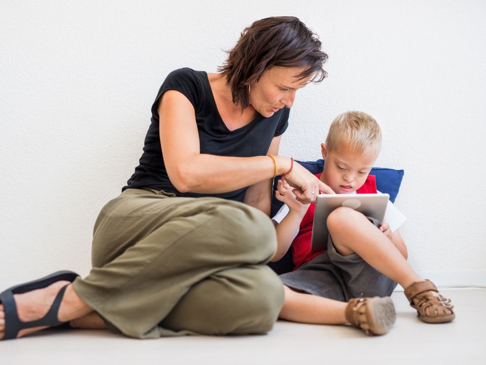 A mother and son sit on the floor, against a wall. The boy, who has Down's syndrome, is looking at a tablet. The mother is pointing to something on the tablet.