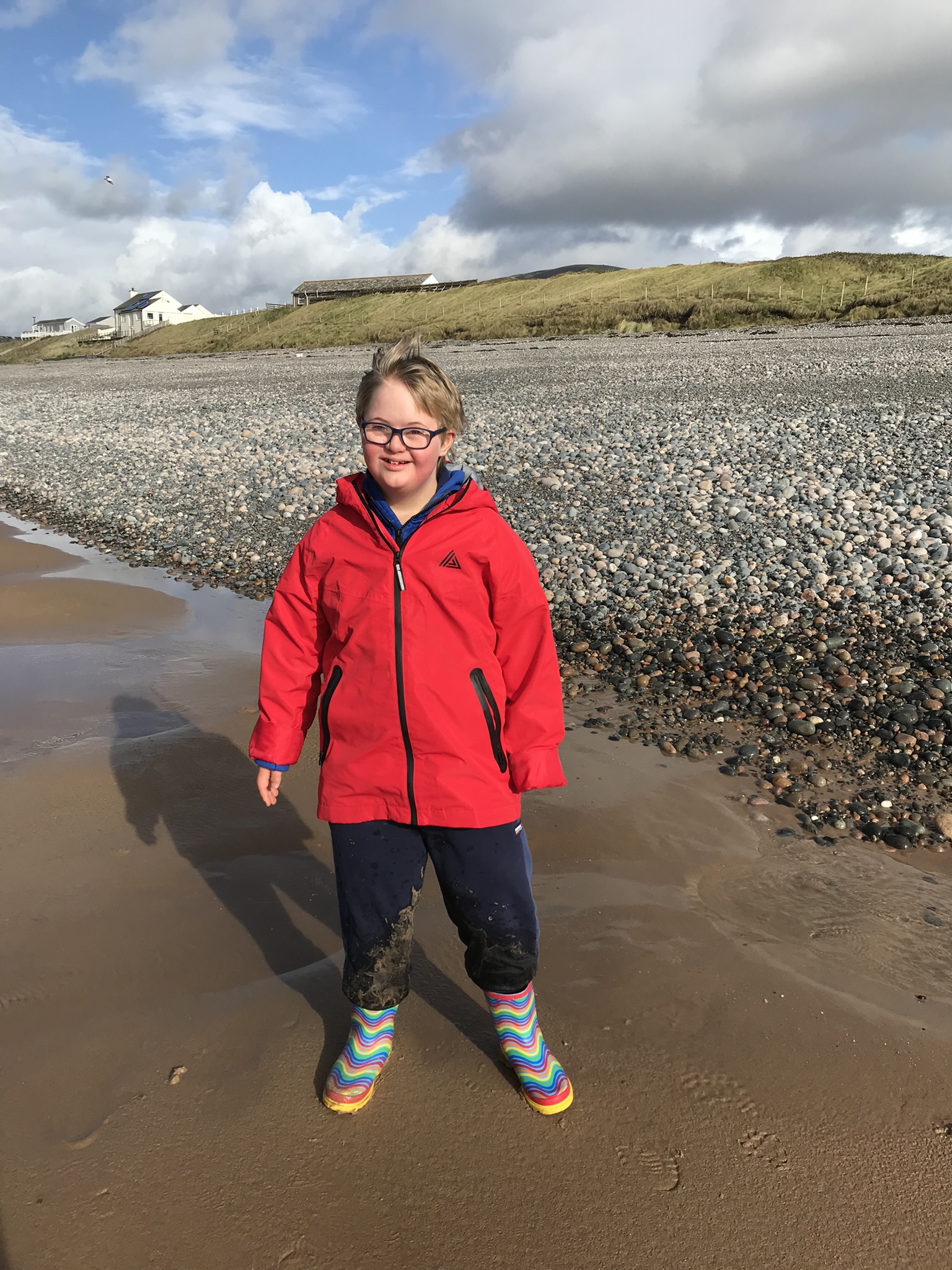 A boy stands on a pebbly beach in a bright red jacket and patterned wellington boots. He is smiling and looking at the camera.