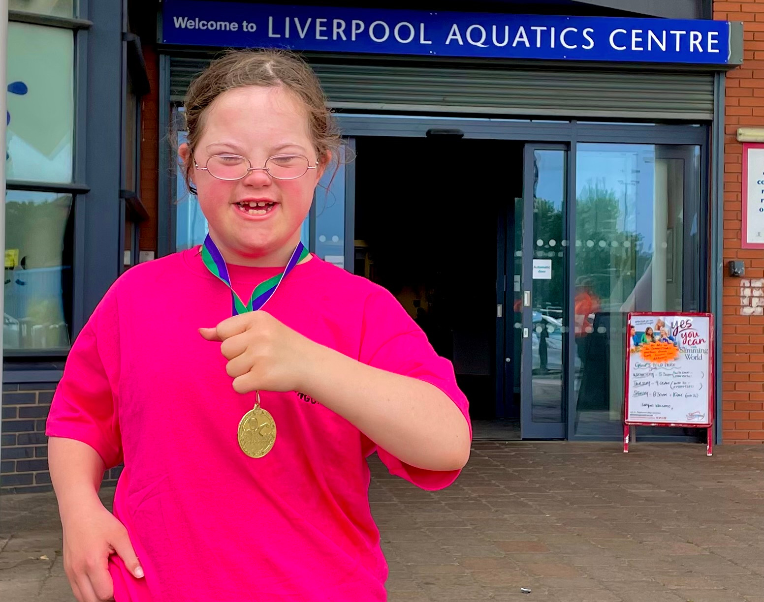 A young girl in a bright pink tshirt stands outside the Liverpool Acquatics Centre. She is smiling broadly and in her left hand she's holding the ribbon to show her gold medal