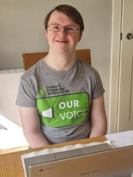 A young man sits at a desk with a laptop and papers in front of him. He is wearing a tshirt with the Our Voice logo on it and wears glasses.