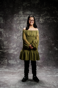A studio photo of Harshi. She is wearing an olive green dress and jeans. She has long, dark hair and glasses and is smiling.