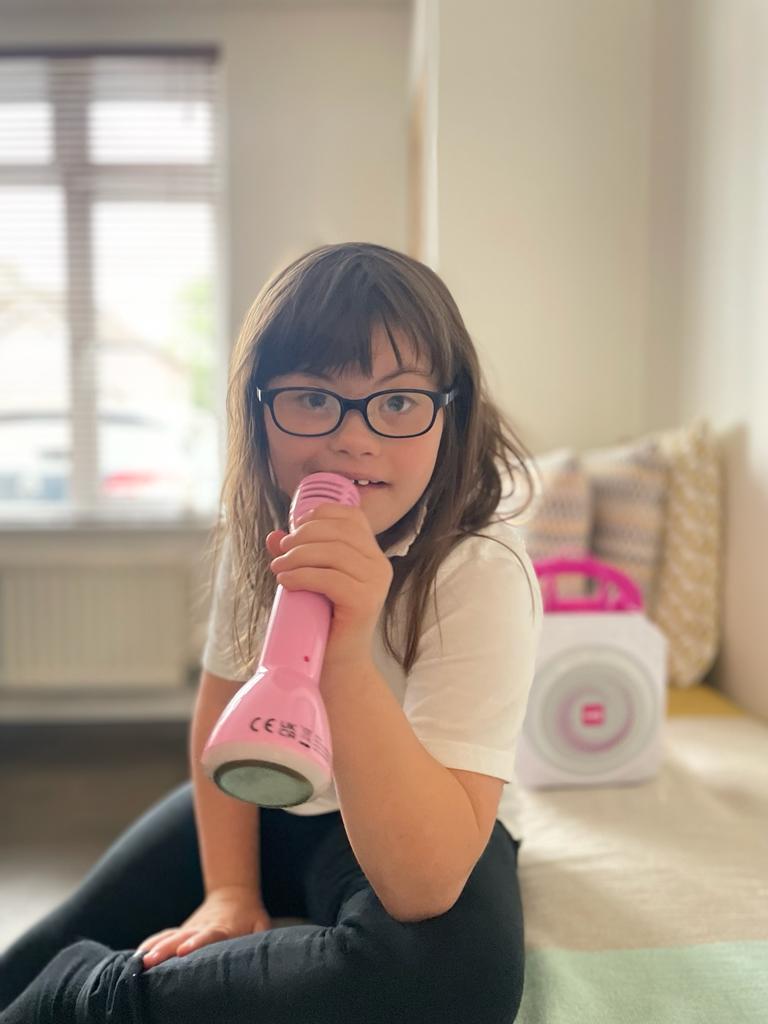 young girl with Down's syndrome is singing into a pink microphone. She is wearing glasses and looking into the camera.
