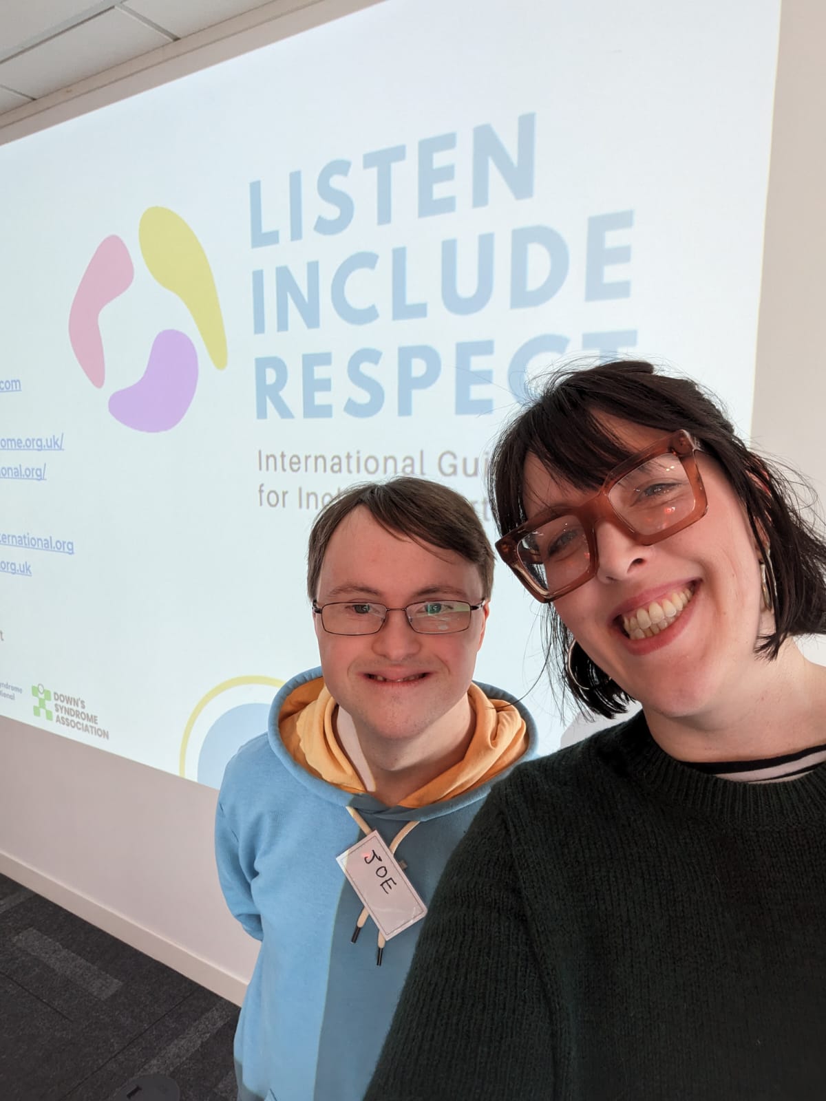 Two people smiling in front of a Listen, Include, Respect backdrop