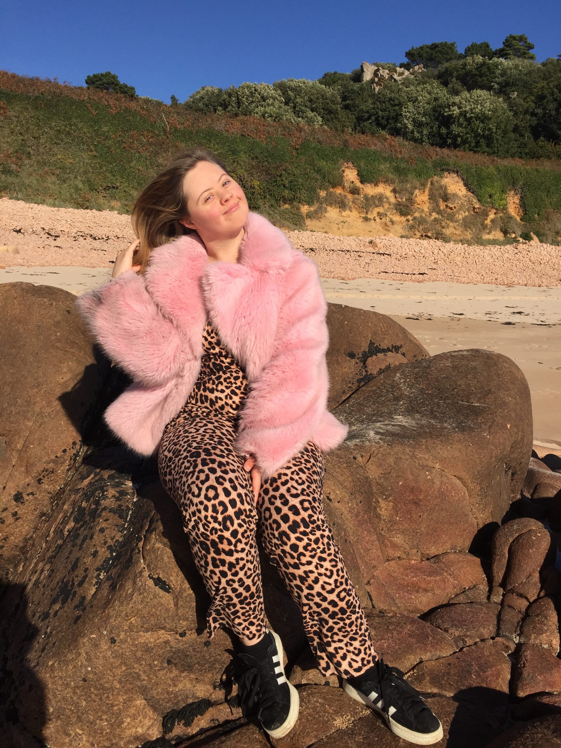 Young woman dressed in leopard print trousers and a pink fur coat, sits on a rock on a beach