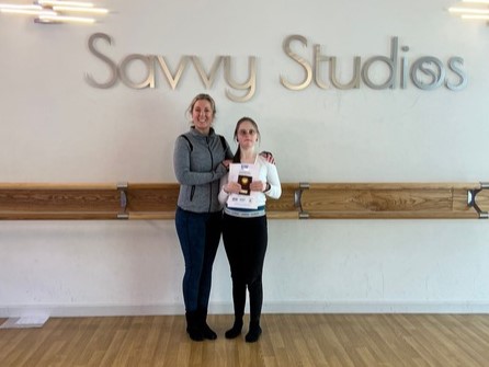 Girls and her ballet teacher stand in front of a Savvy Studios sign holding a certificate